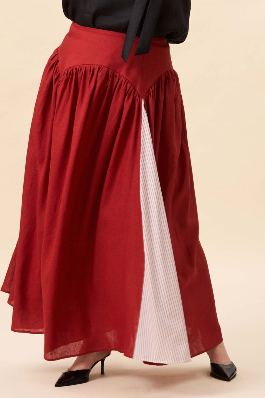 Cordelia Gray maxi skirt in tomato red  The waist holds a fitted structure before releasing into a full, gathered skirt. Contrasting panels at the front and back shape the silhouette, introducing variation and rhythm as the garment moves.