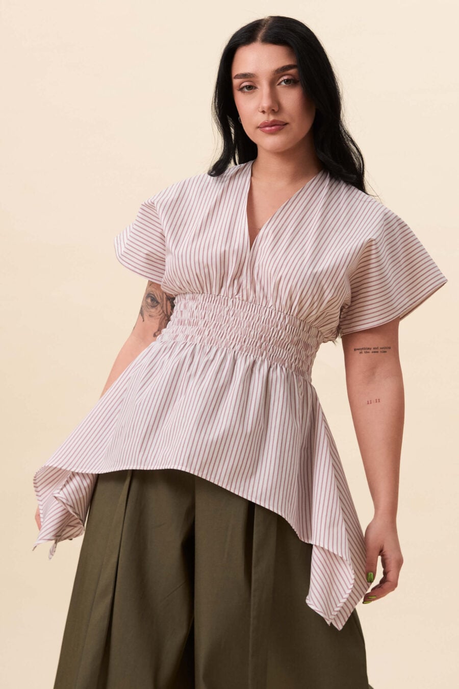 Nemesis ruched top in red stripes - A structured top defined by a deep V neckline and a shaped waist panel. The bodice is fitted through a smocked section, creating tension and adaptability. From the waist, the fabric releases into soft, pointed extensions that move away from the body.