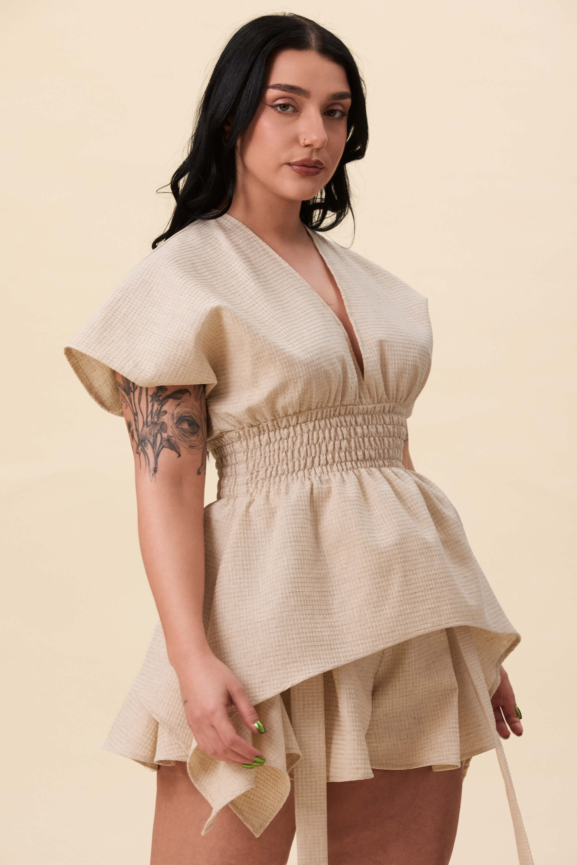 Spring 2026 Nemesis Ruched Top In Textured Beige 1 Nemesis Ruched Top In Textured Beige Nemesis ruched top in textured beige A structured top defined by a deep V neckline and a shaped waist panel. The bodice is fitted through a smocked section, creating tension and adaptability. From the waist, the fabric releases into soft, pointed extensions that move away from the body.