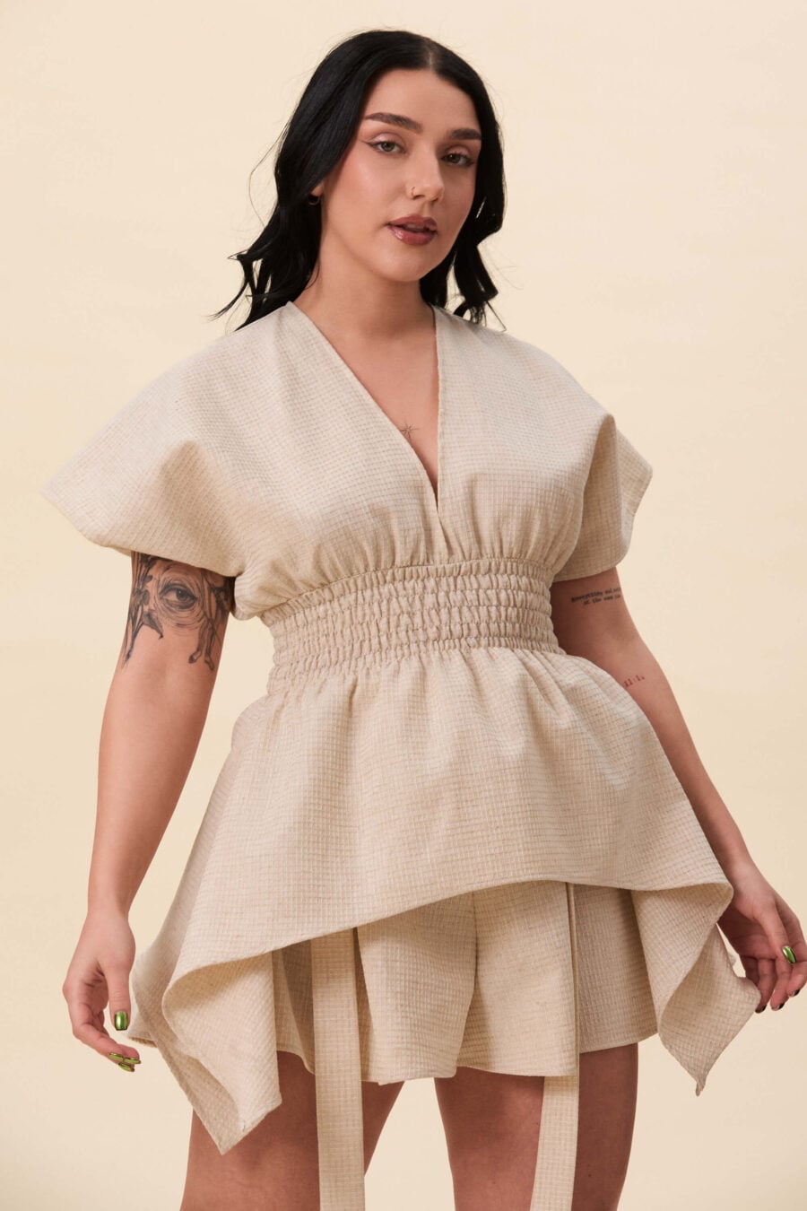 Nemesis ruched top in textured beige - A structured top defined by a deep V neckline and a shaped waist panel. The bodice is fitted through a smocked section, creating tension and adaptability. From the waist, the fabric releases into soft, pointed extensions that move away from the body.